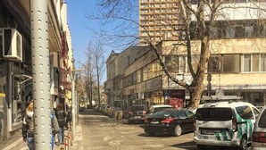 A street scene with parked cars, a building with a sign, and a tree. - Central Studio 2 (Burgas)