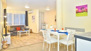 A modern living room with a sofa, dining table, and chairs. There is a kitchen area with a refrigerator and a dining table set with plates and glasses. - Central Studio 2 (Burgas)