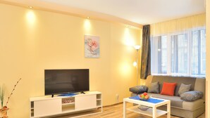 A living room with a flat-screen TV on a white stand, a sofa with cushions, a small table with fruit, and a window with curtains. - Central Studio 2 (Burgas)
