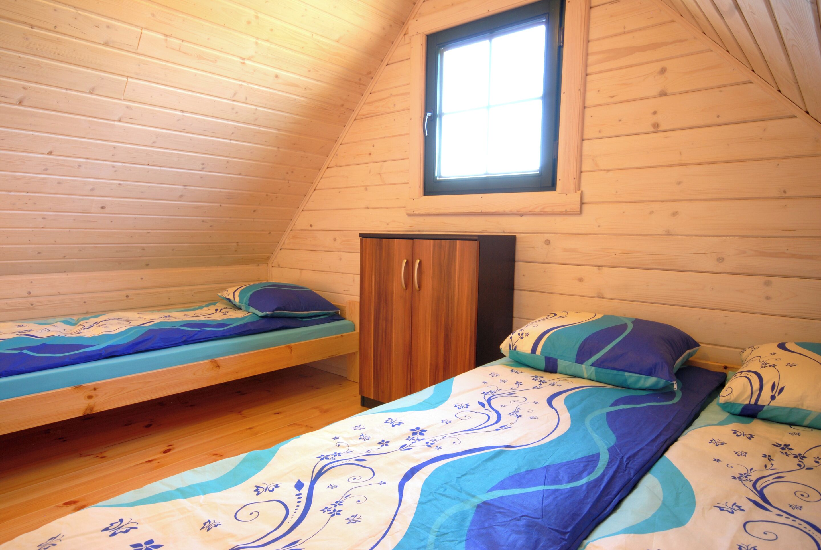 A small, cozy room with wooden walls and a sloped ceiling. It contains two single beds with matching blue and white bedding, a wooden wardrobe, and a window providing natural light.