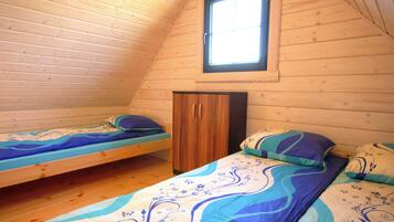 A small, cozy room with wooden walls and a sloped ceiling. It contains two single beds with matching blue and white bedding, a wooden wardrobe, and a window providing natural light.