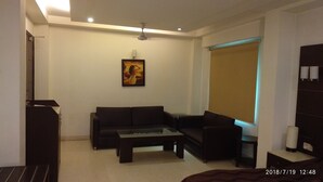 A living room with a sofa, a coffee table, a painting on the wall, and a window with a blind. - The Metropolis Guest House Near RK Ashram Metro Station (New Delhi)