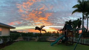 A playground with a slide and climbing structure, surrounded by a fenced yard with grass and palm trees. - Cozy Tropical Retreat Guest House with AC (Hilo)