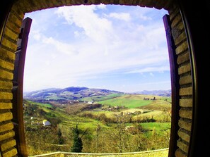 A view through a window with stone framing, overlooking a rural landscape with rolling hills and scattered buildings. - Nidastore B&B (Arcevia)