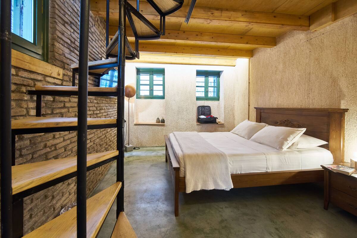 Quarto com cama de madeira, mesa de cabeceira, luminária, parede de tijolos e janela com persianas verdes.