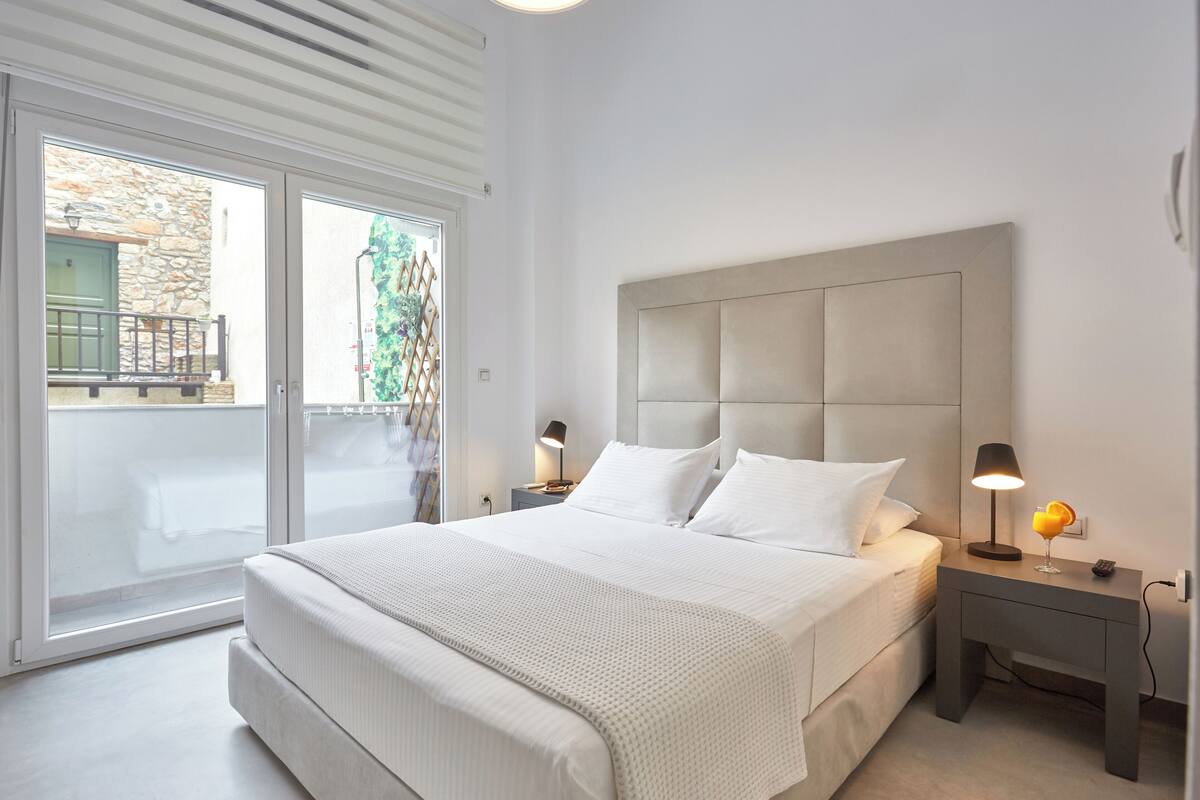 Quarto moderno com uma cama grande, mesa de cabeceira com abajur e vista para o exterior por uma porta de correr.