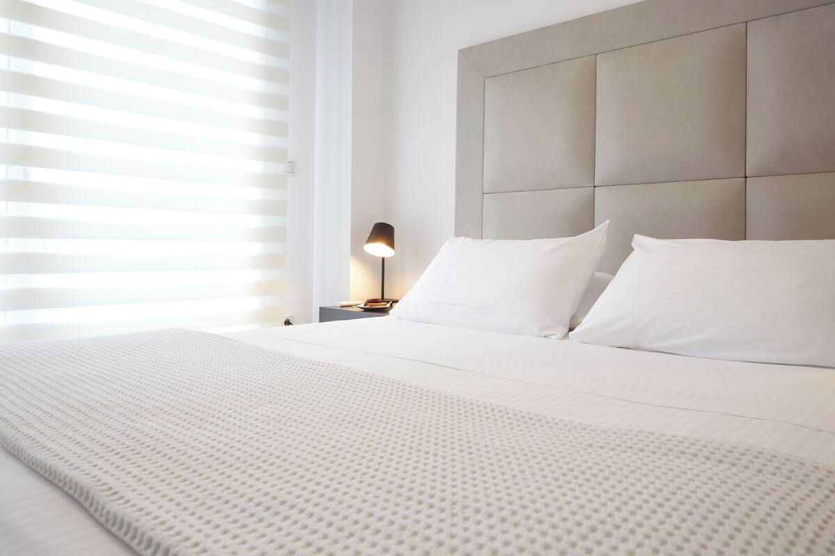 Uma cama bem arrumada com cobertura branca texturizada, cabeceira com design acolchoado e luminária de mesa em um criado-mudo.