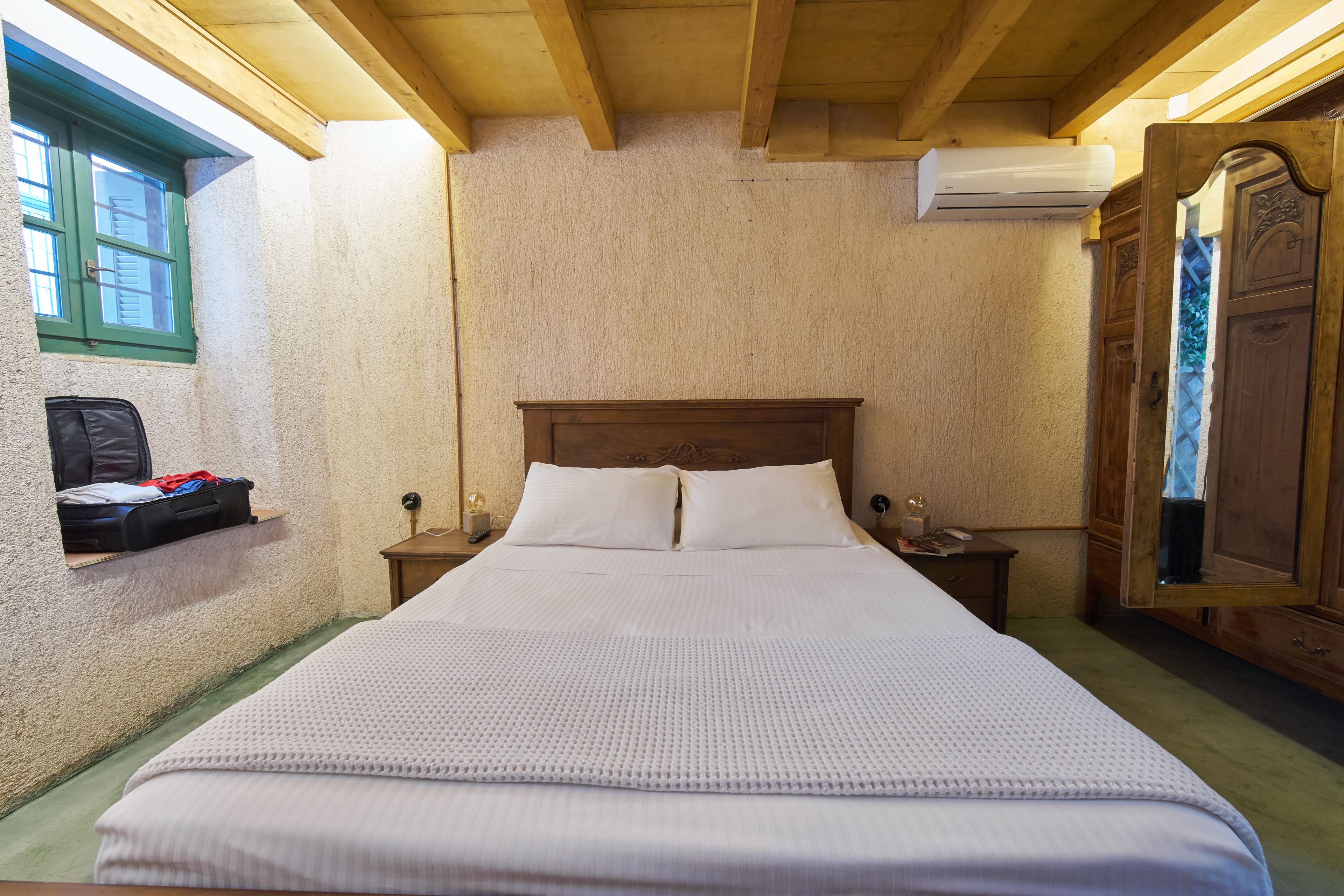 Una cama bien hecha en una habitación con techo de madera, una mesita de noche pequeña con una lámpara, un aire acondicionado instalado en la pared y una ventana con marco verde.