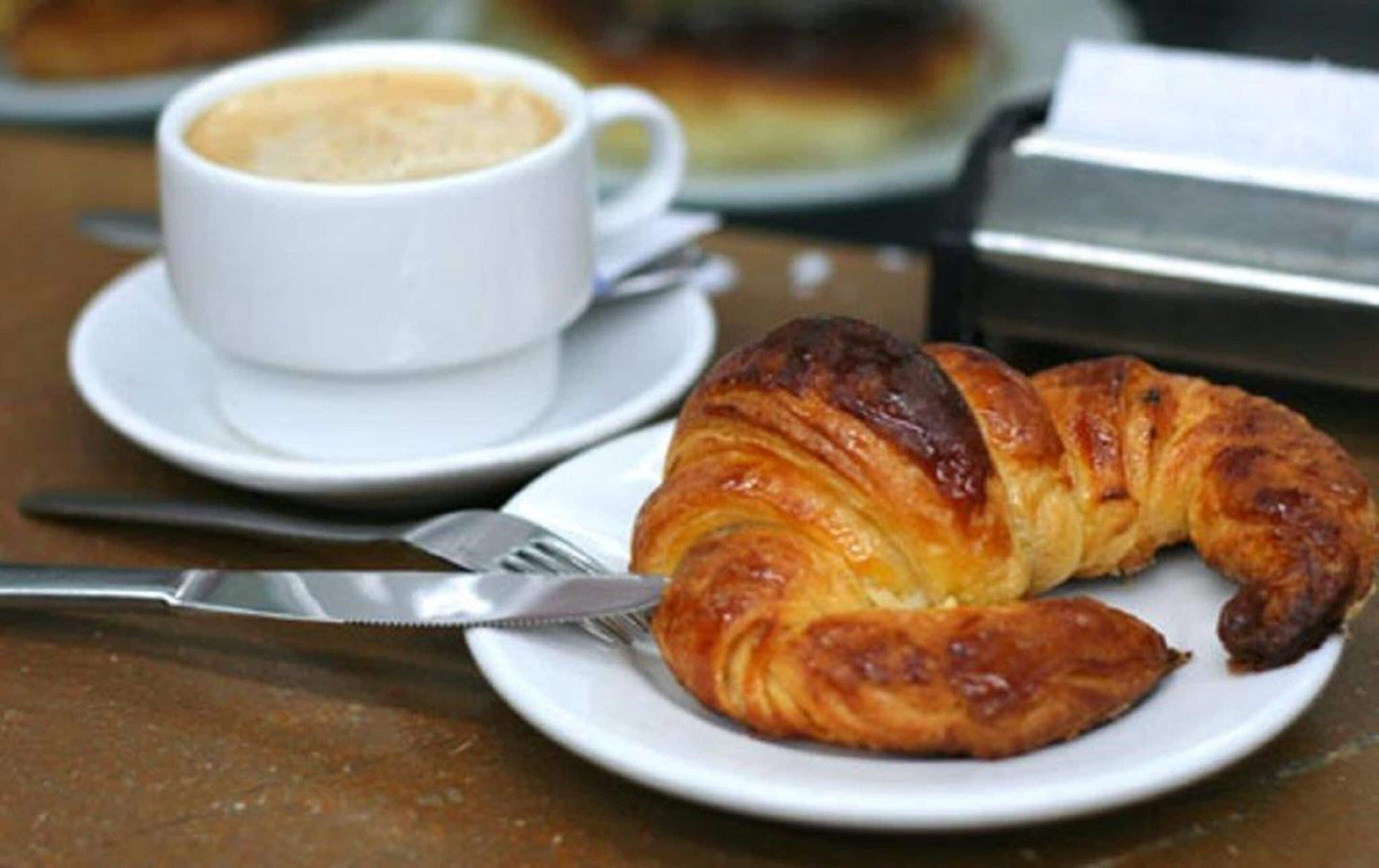 A croissant on a plate with a cup of coffee and a knife on a table.