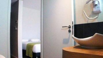 A hotel room with a bed, a wooden bedside table, and a modern bathroom with a large sink and a hairdryer mounted on the wall.