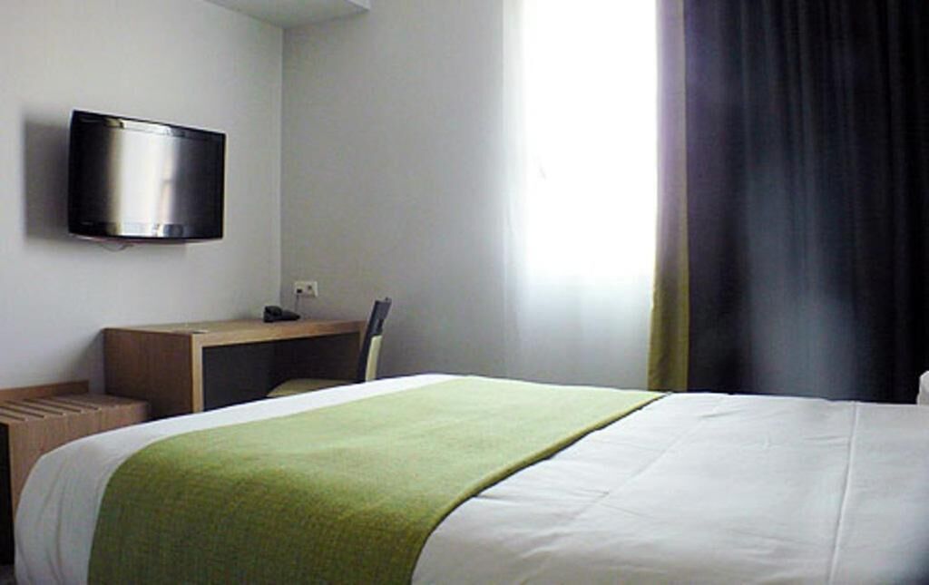 Et hotelværelse med seng, skrivebord, stol og et fjernsyn monteret på væggen.