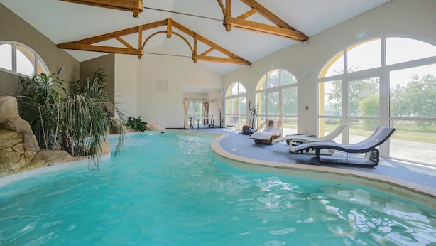 An indoor swimming pool with clear blue water, wooden beams on the ceiling, and large windows offering views of the outdoors.