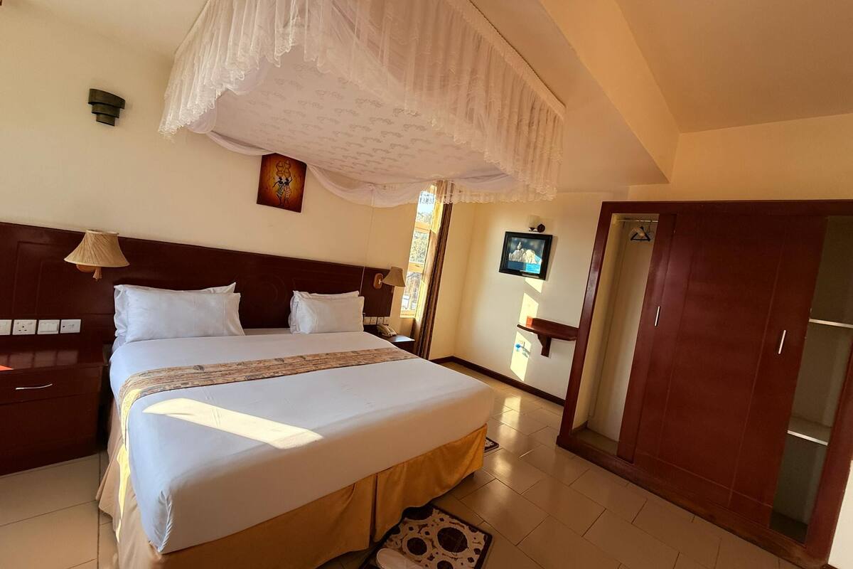 A hotel room with a large bed, a wooden headboard, a bedside table with a lamp, a TV mounted on the wall, and a wardrobe.