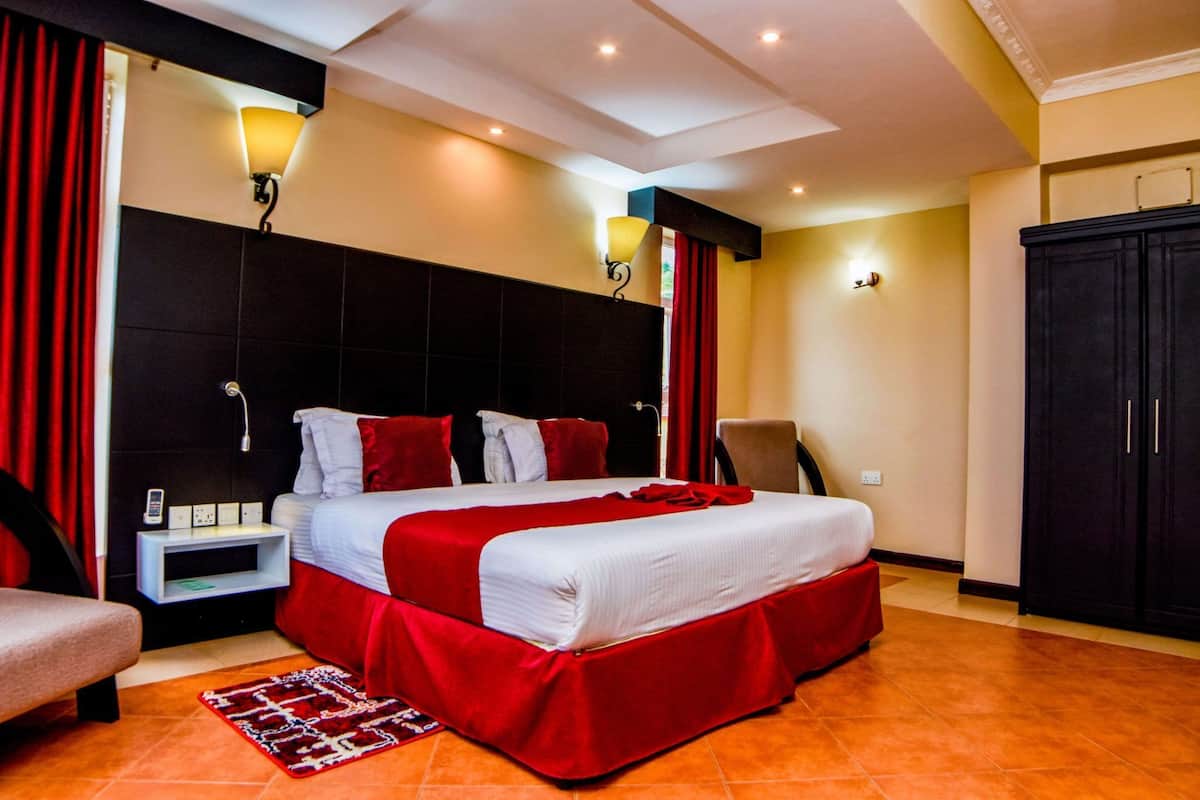 A hotel room with a large bed, red curtains, a nightstand, a chair, and a wardrobe.
