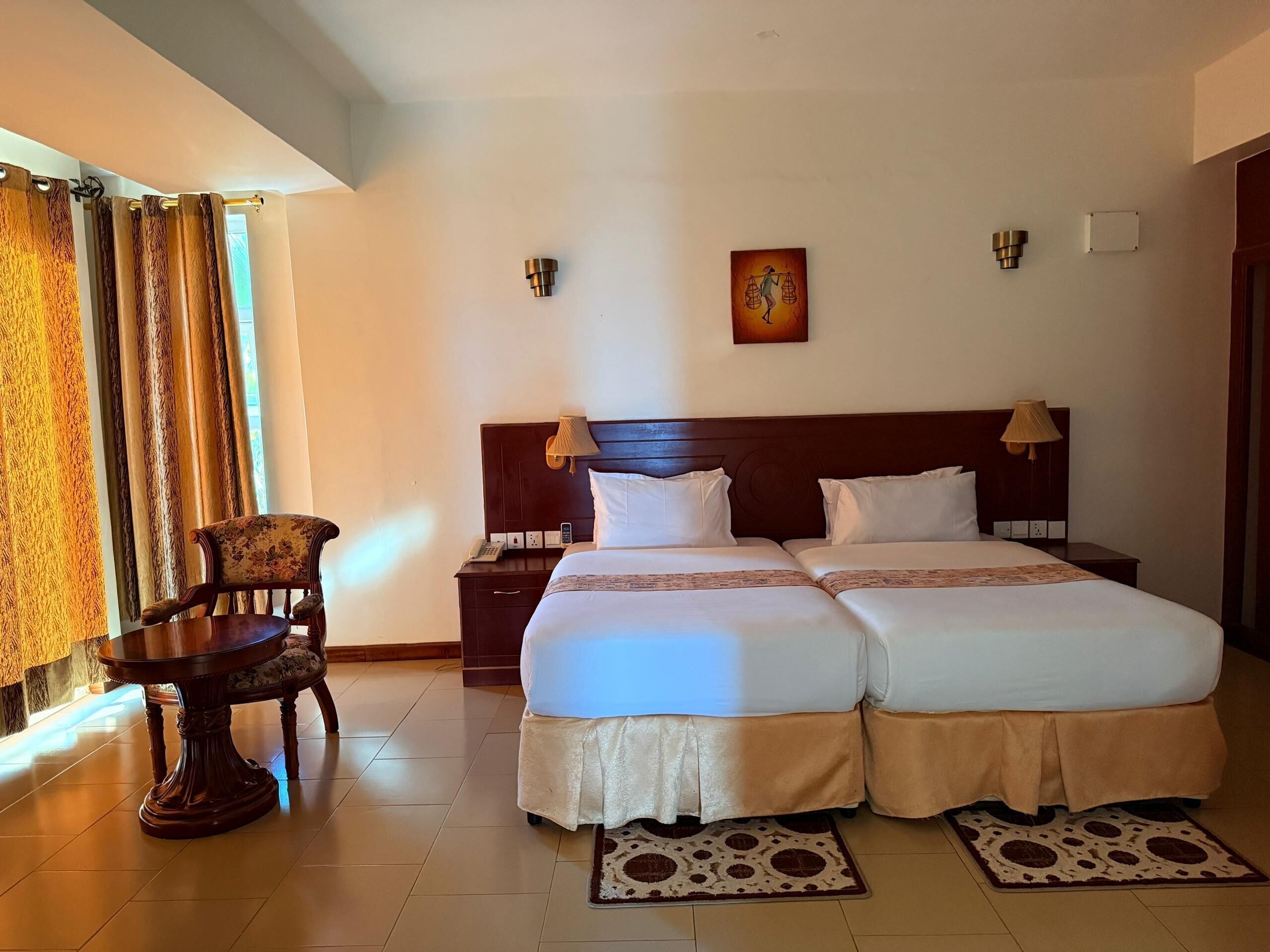 Quarto de hotel com duas camas, uma cadeira de madeira, uma mesinha e um quadro na parede.
