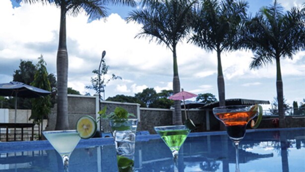 A poolside area with a glass table displaying various drinks, palm trees, and a cloudy sky. - ISAMILO GRAND HOTEL (Mwanza)