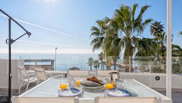 A table set for breakfast with croissants, orange juice, and coffee, overlooking a beach with palm trees and the sea.