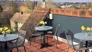 A rooftop terrace with tiled flooring, metal chairs, and round tables, each with a vase of yellow flowers. - The Black Hole (Winchester)