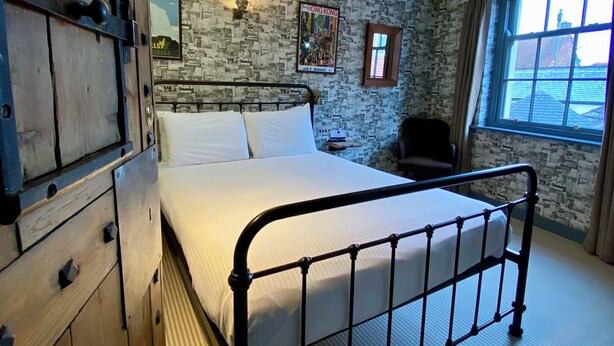 A bedroom with a metal bed frame, a stone-patterned wall, a window with curtains, and a mounted fire alarm. - The Black Hole (Winchester)