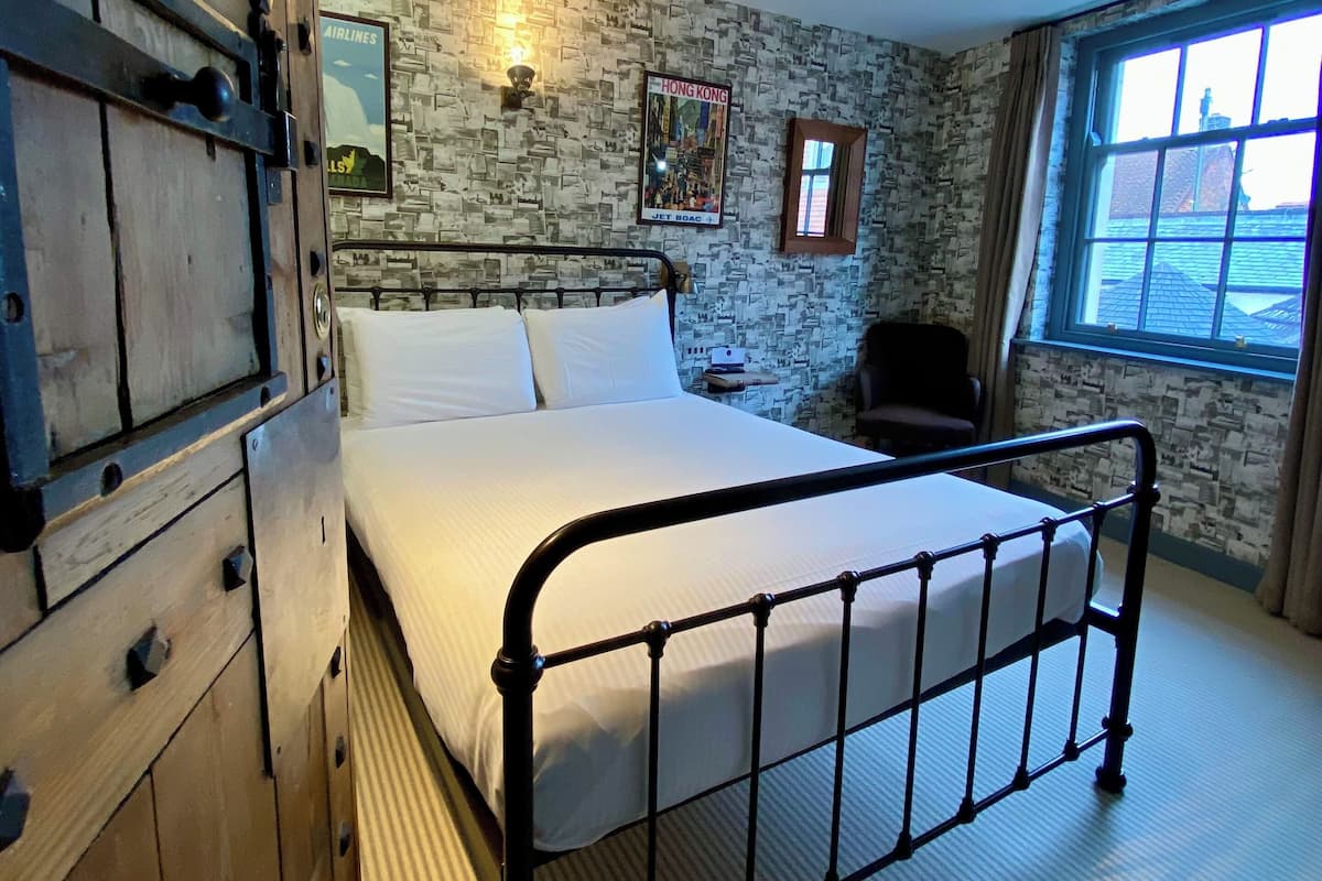 A bedroom with a metal bed frame, a stone-patterned wall, a window with curtains, and a mounted fire alarm.