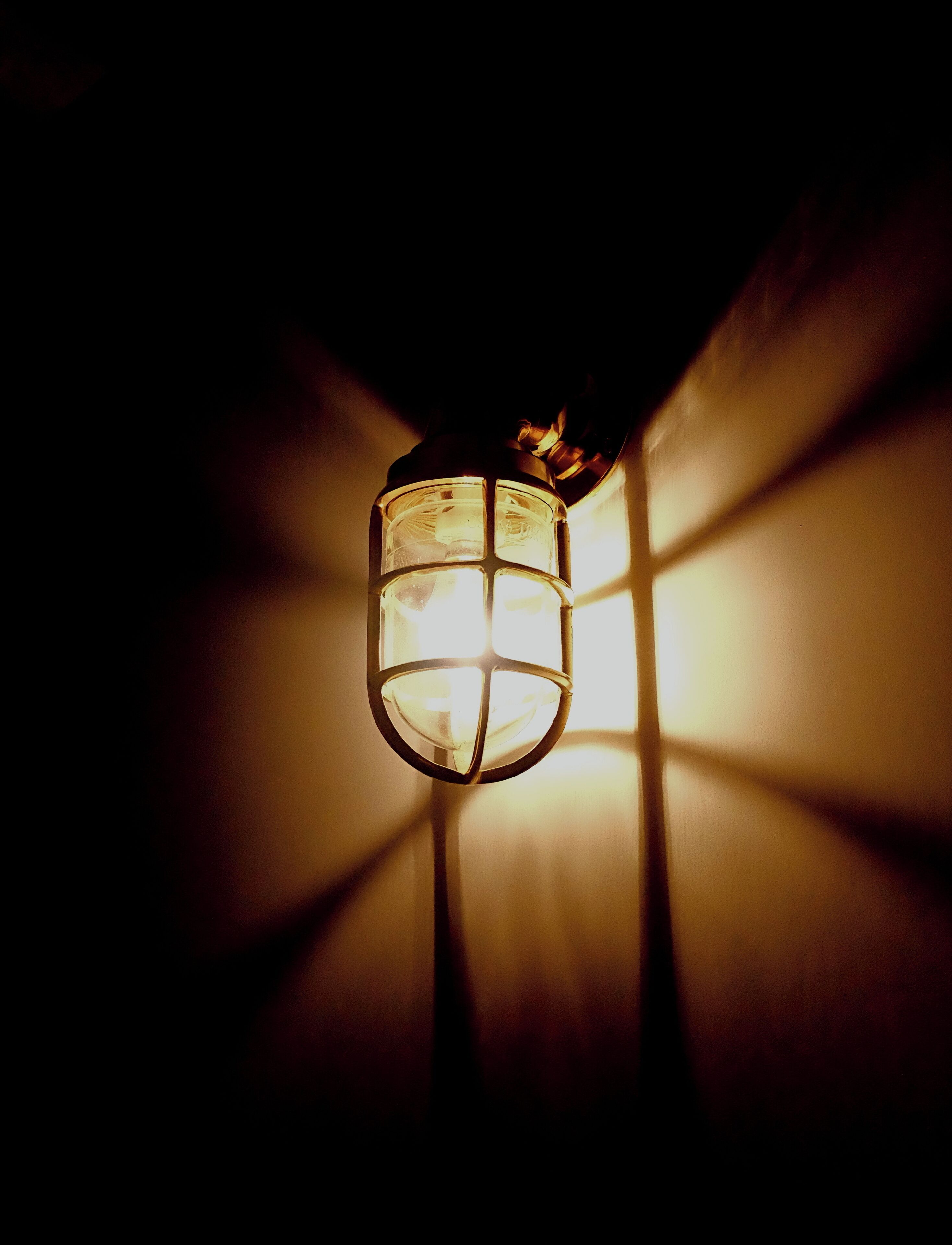 Un farol colgante con una cubierta de vidrio y estructura de metal, proyectando luz sobre una pared.