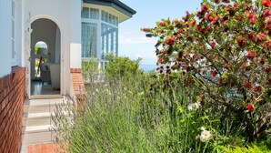 Une maison moderne dotée d’une porte vitrée donnant sur un porche abrité, le tout entouré d’une végétation luxuriante et de plantes à fleurs.