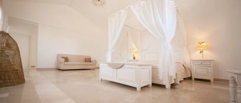 A bedroom with a four-poster bed, a bedside table with a lamp, a nightstand, a sofa, and a wicker chair.