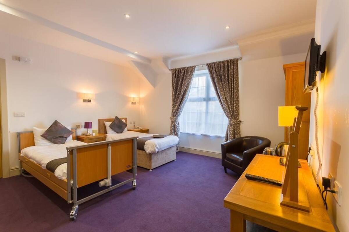 Habitación de hotel con cama, mesita de noche, una silla, escritorio, lámpara, televisor y ventana con cortinas.