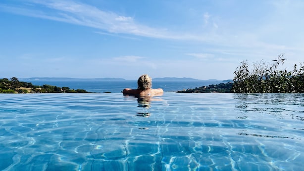 Una persona relajándose en una piscina infinita transparente con vistas al mar y montañas distantes.