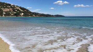 Una playa con agua cristalina, una orilla arenosa y una colina con edificios al fondo.