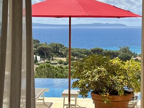 A view of a swimming pool with a red umbrella, a potted plant, and a scenic landscape including trees and the sea. - Villa Thalassa Art' B&B (Le Lavandou)