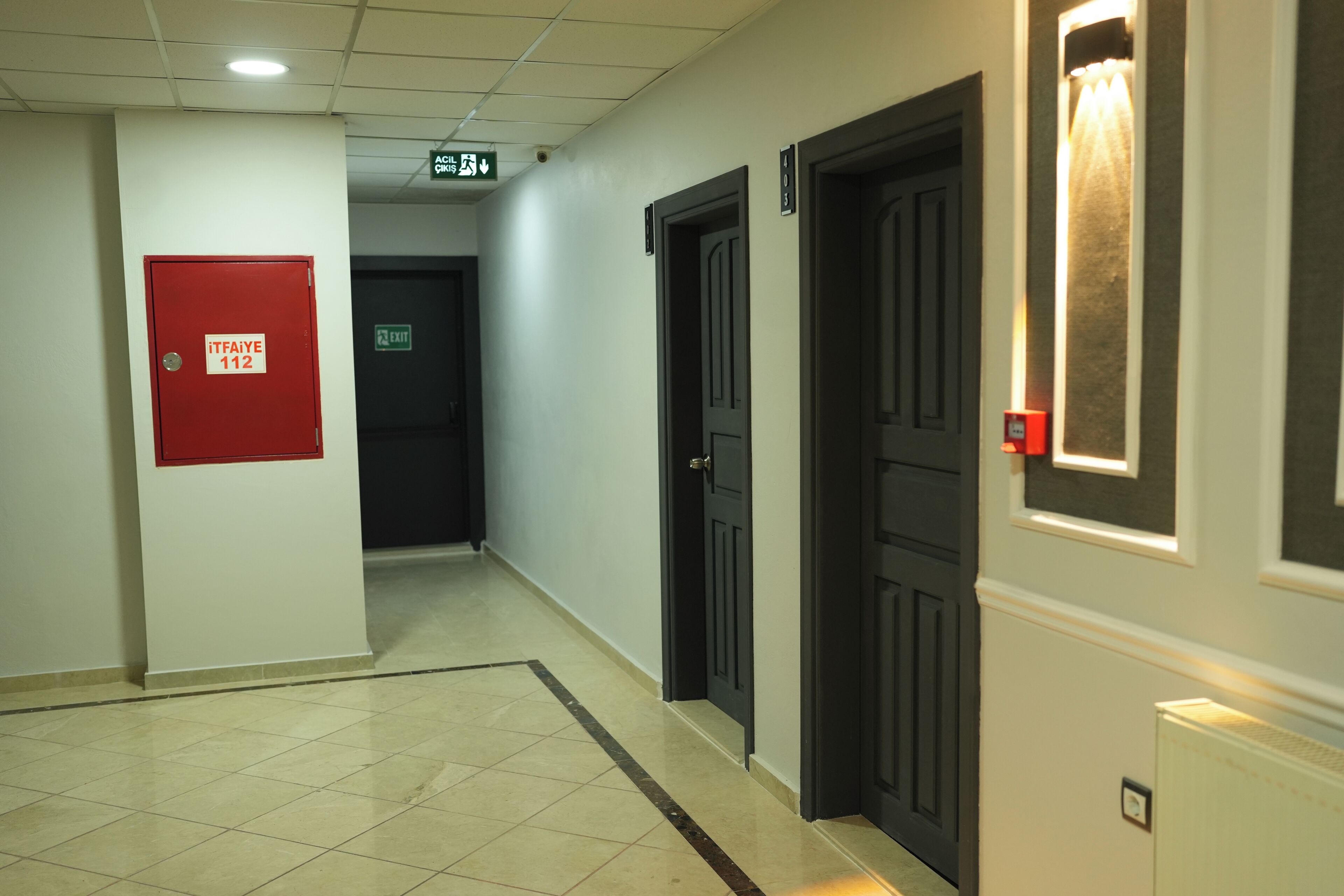 A hallway with a red emergency exit door labeled "itfaiye 112", black doors, and a tiled floor.