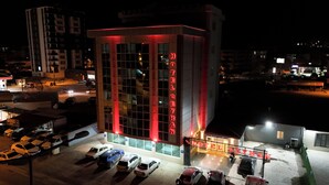Un hôtel reconnaissable à son grand panneau néon rouge, un parking où sont garées plusieurs voitures, et une vue sur le paysage urbain environnant la nuit.