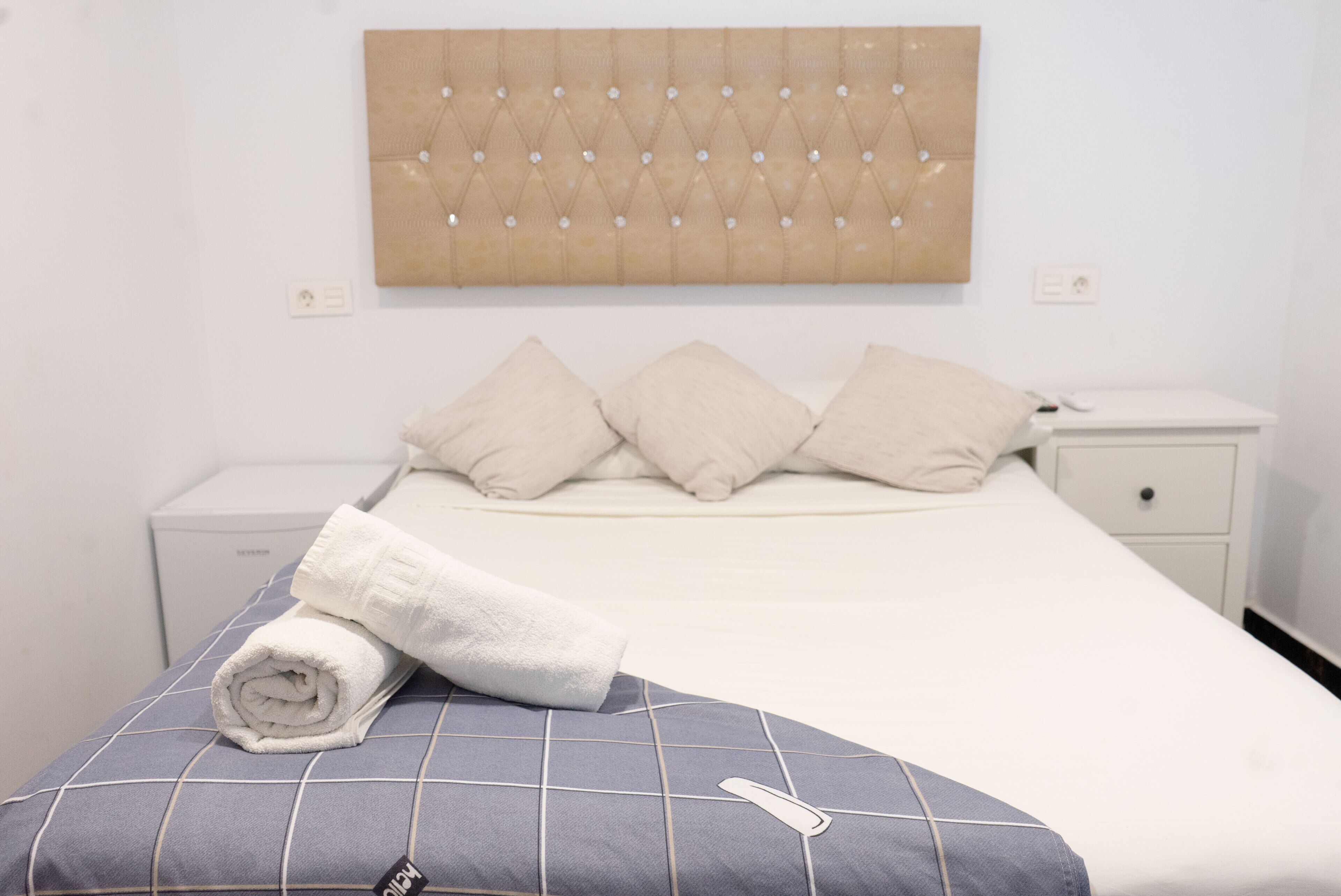 Una cama bien tendida con una toalla blanca enrollada, un cabecero capitonado y una mesita de noche con cajón.