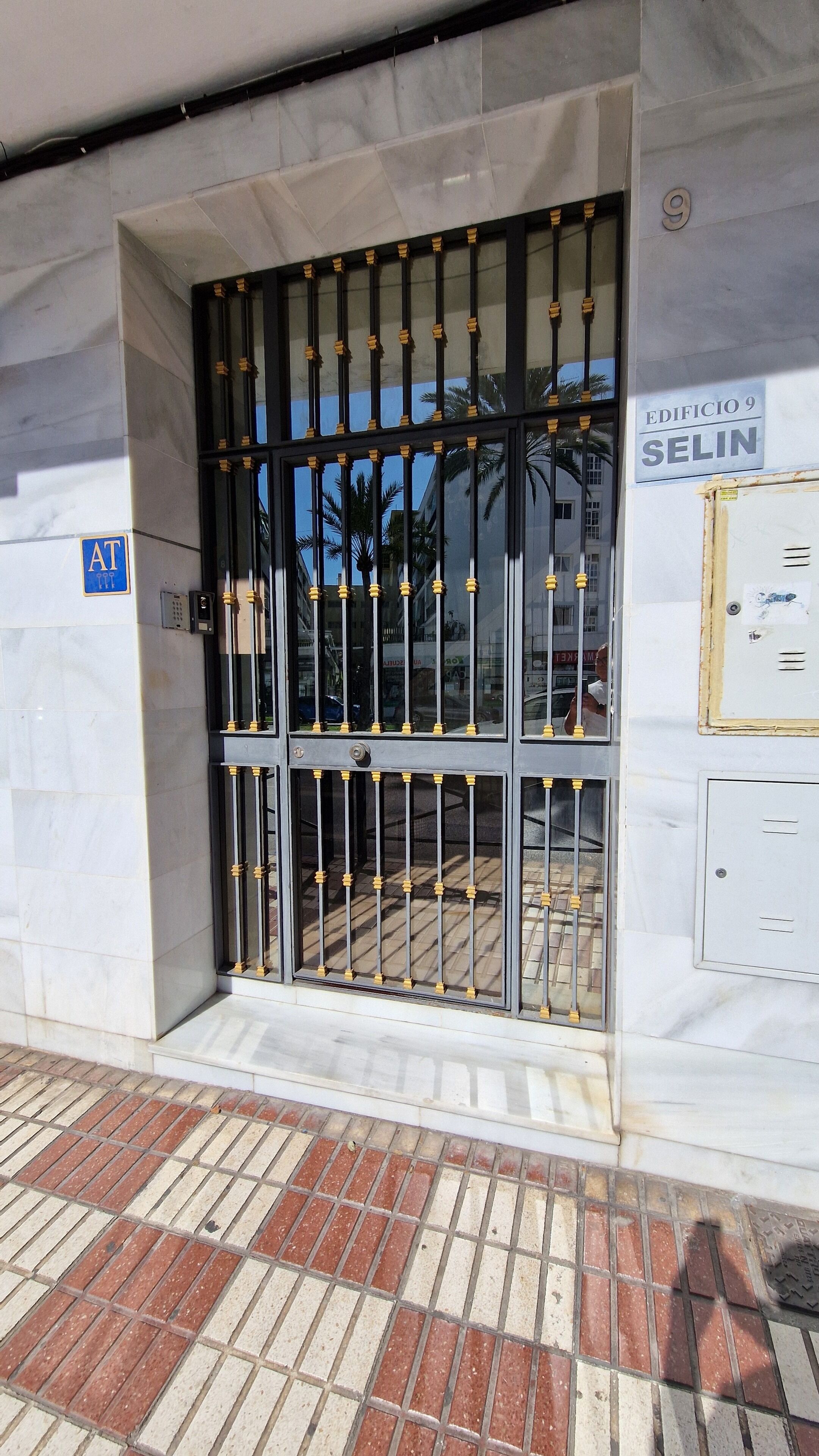 Entrada de un edificio con una reja metálica, un cartel azul con el símbolo de AT y un letrero que dice 'EDIFICIO 9 SELIN'.