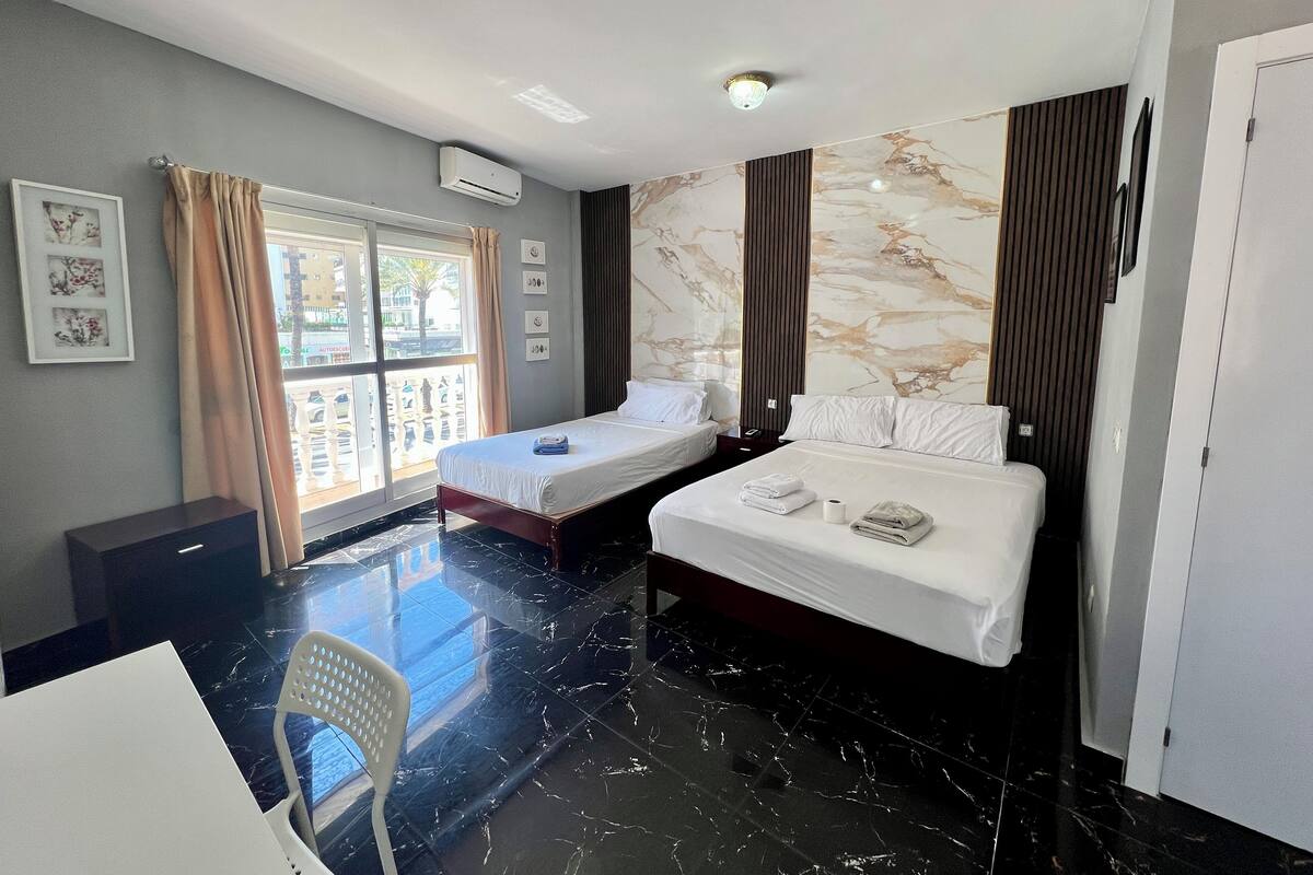 A hotel room with two beds, a large window with curtains, a marble-patterned wall, and a ceiling light.