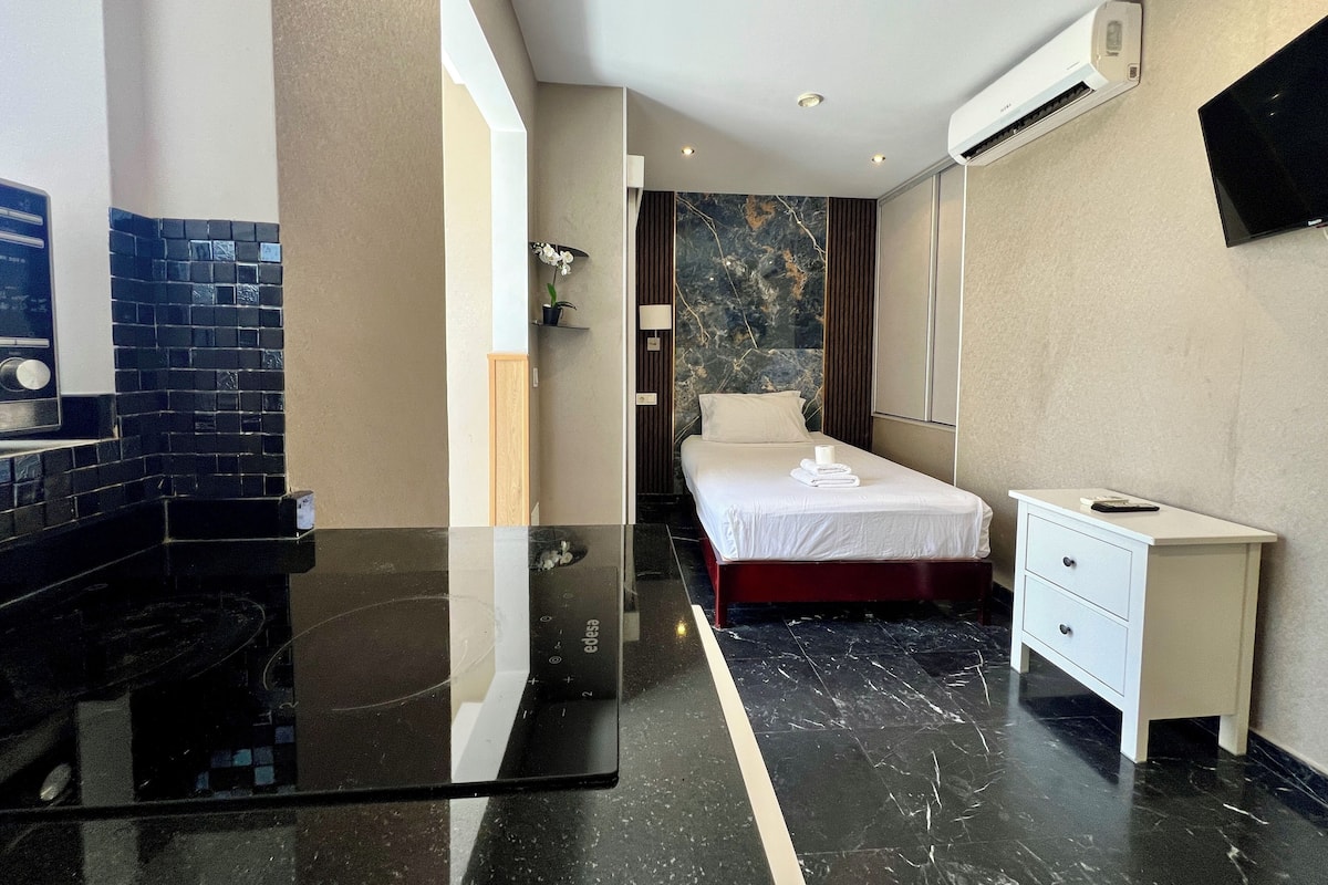 A hotel room with a bed, a bedside table, a desk, a mirror, an air conditioning unit, and a TV mounted on the wall.