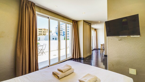 Habitación de hotel con una cama grande, televisor de pantalla plana montado en la pared y puerta corrediza de vidrio que da a un área exterior con una mesa y sillas.