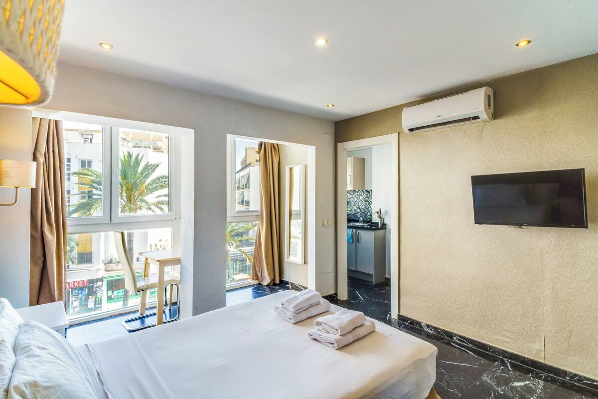 A hotel room with a bed, a TV, an air conditioning unit, and a view of the outside.