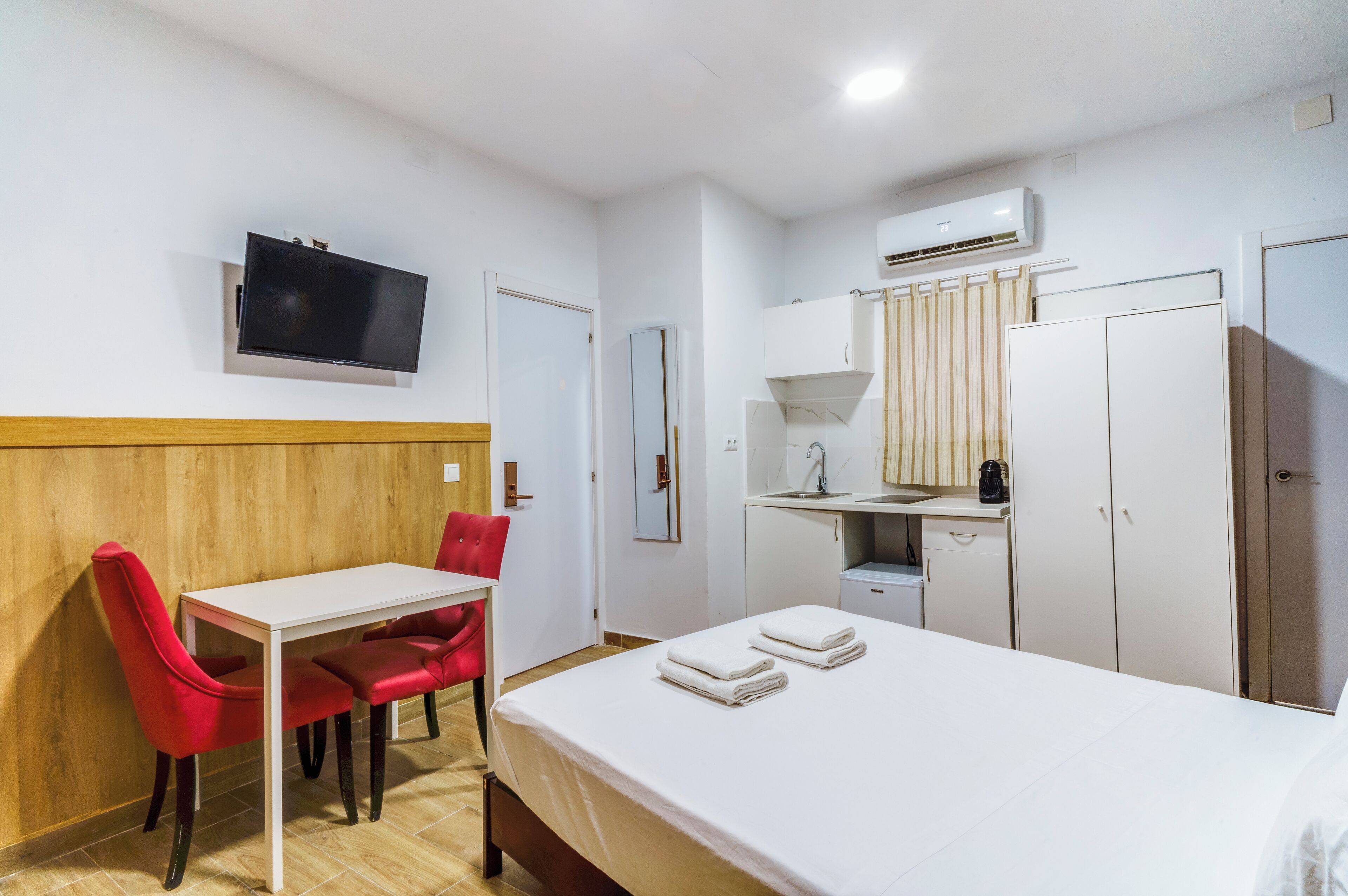 Habitación de hotel con cama, escritorio y silla, televisor montado en la pared, armario y una kitchenette pequeña.
