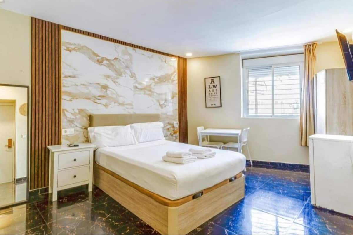 A hotel room with a bed, bedside table, a desk, and a window.