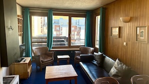 A cozy room with a wooden floor, a sofa, a table, and a chair. There are windows with curtains, and a view of buildings outside.