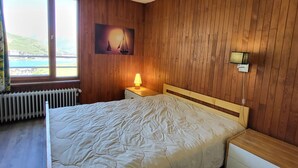 Ein Schlafzimmer mit einer holzverkleideten Wand, einem Bett mit weißer Steudecke, einem Nachttisch mit Lampe, einem Gemälde mit Segelbooten und einem Fenster mit Meerblick.