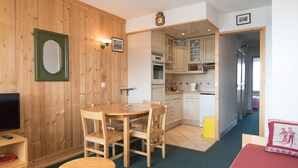 Une pièce chaleureuse aux murs en bois, avec une table à manger et des chaises, une petite table ronde, un lit recouvert d’une couverture rouge, et un coin cuisine équipé de placards et d’un évier.