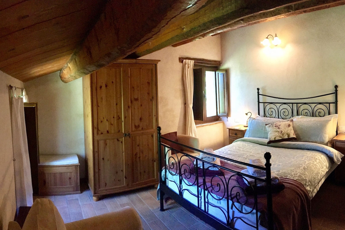 Een slaapkamer met een houten plafond, een bed met een metalen hoofdbord, een nachtkastje, een kledingkast en een raam met gordijnen.
