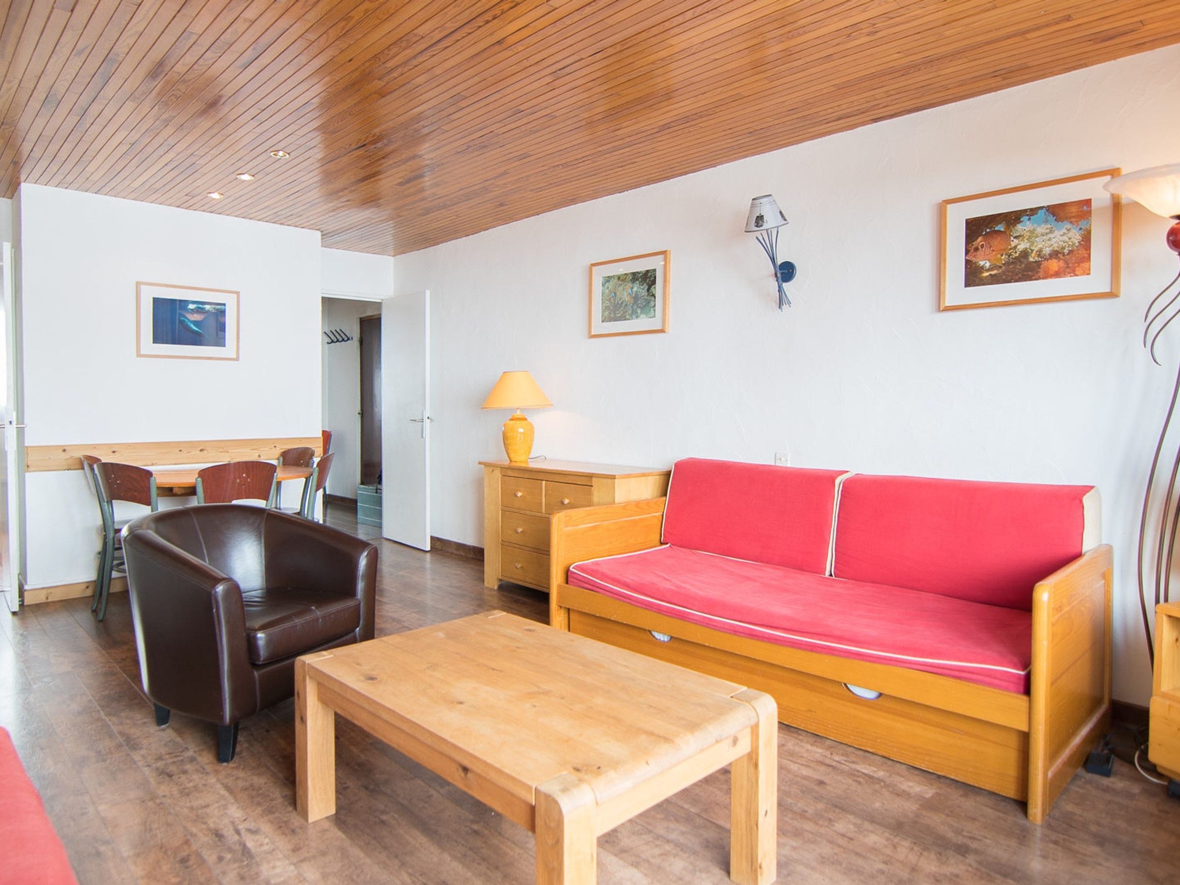 A room with a wooden ceiling, a red sofa, a wooden table, a lamp, and a painting on the wall.