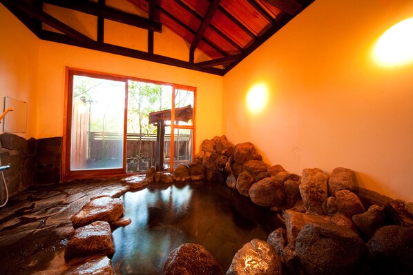 Un onsen japonais traditionnel, avec un bain en pierre entouré de rochers, des poutres en bois au plafond et une porte en bois donnant sur l’extérieur.