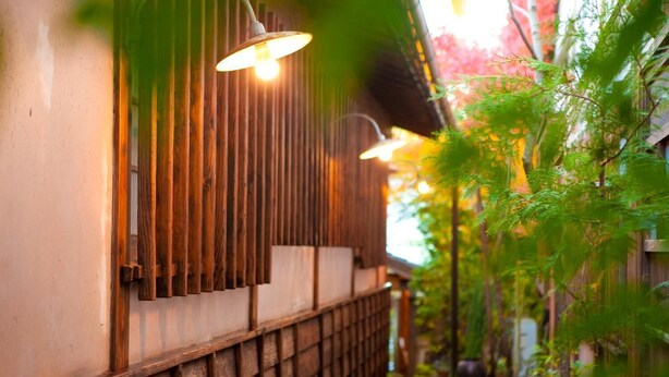 A narrow pathway flanked by wooden fences and green foliage, leading to a building with vertical wooden slats and hanging lights. - Yufuin TAGASODE (Yufu)