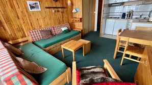 A cozy cabin interior with a wooden wall, a sofa with plaid cushions, a small table, and a dining area with a wooden table and chairs.