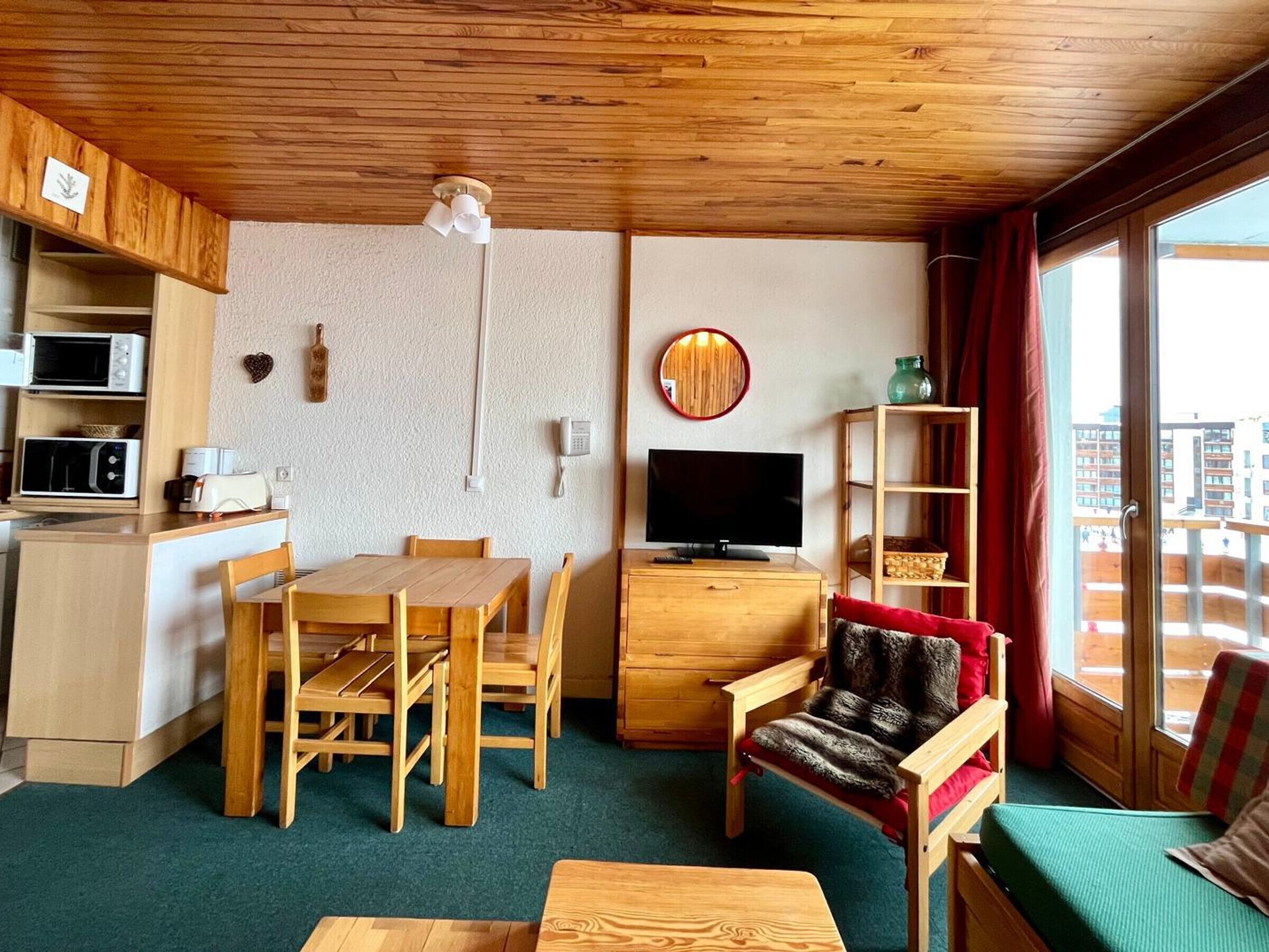A compact living space with a wooden ceiling, a dining table with chairs, a sofa, a television, and a small kitchen area with a microwave and toaster.